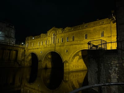 The image displays Pulteney Bridge in Bath, United Kingdom, at night. The multi-arched stone bridge is topped with continuous Georgian-style buildings, all strongly illuminated by a warm yellow artificial light. This illumination highlights the architectural details, including a central classical facade with a prominent arched window and pediment, and numerous rectangular windows on the building's two visible levels. The warm light creates elongated reflections on the calm, dark water of the river flowing beneath the bridge. To the right, a dark stone retaining wall is partially visible, topped by a dark metal railing system. A large stone building with a rounded roof section is partially discernible in the background on the far left. No individuals or discernible human activity are present in the scene.