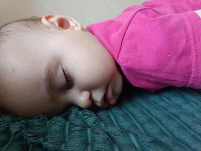 The image is a close-up, eye-level shot of a young child sleeping. The child is lying on their side, with their head resting on a textured, teal-colored blanket. The child has fair skin and dark eyelashes. They are wearing a bright pink t-shirt. The background is a plain, light-colored wall. There is no indication of the child's location in the city of Посад, Russia, or any other specific location. The scene is peaceful and intimate, focusing solely on the sleeping child.