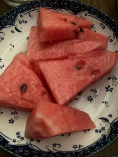 This media file is a close-up, overhead shot of several triangular slices of watermelon arranged on a white plate with a blue floral pattern. The watermelon slices are a vibrant pink and appear juicy. Some of the slices have visible black seeds. The plate itself has a dark blue rim with a repeating pattern of small blue and white flowers. The background is blurred, but appears to be a wooden surface. There is no visible text in the image. The lighting suggests it might be daytime. The scene depicts a simple still life of fruit.