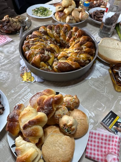 The image shows a table laden with various food items, seemingly at a gathering in Vinarovo, Bulgaria. A large twisted loaf, topped with almonds, sits in a grey pan. Next to it is a white plate piled high with an assortment of baked goods, including small rolls with colorful sprinkles, rounded cookies, and croissant-shaped pastries dusted with sugar. Other dishes on the table contain salads, sliced bread, and meat dishes. A pack of cigarettes and a checkered napkin lie nearby. A bottle of liquid is also visible. The tablecloth has a faint pattern of culinary-related imagery and text.