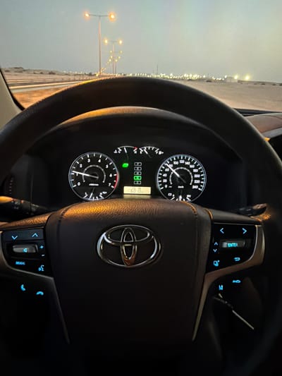 The image shows the interior of a car, specifically the dashboard and steering wheel, as the vehicle is moving along a road at night in Umm Salal Ali, Qatar. The dashboard displays a speedometer reading around 60 km/h and an odometer displaying 88888 km. The steering wheel is centered, featuring the Toyota logo. The car is on a road with streetlights visible through the windshield on the left side of the frame.