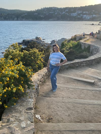 A medium, long-shot outdoor photograph taken during the day features a woman posing on a walkway next to the coast, likely in Palafrugell, Spain. The woman is centered and facing the camera, wearing a light blue and white striped shirt, blue jeans, and a belt. She has one hand on her hip. The walkway is made of concrete steps and a stone wall borders the edge near the water. Yellow flowering bushes grow over the wall. In the background, the Mediterranean Sea is visible, along with a distant shoreline and buildings. In the distance to the right, other people are visible on the walkway. The lighting is bright, suggesting a sunny day.
