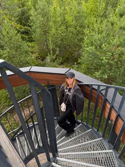 An outdoor, high-angle shot shows a Caucasian woman standing on a metal spiral staircase. She is wearing a black baseball cap, black jacket, black sweatshirt, and black pants. She has a small, white, woven purse with a gold chain strap. The staircase is surrounded by a dark gray metal railing and is set against a backdrop of lush green trees. The location is the Peski, Russia city area.
