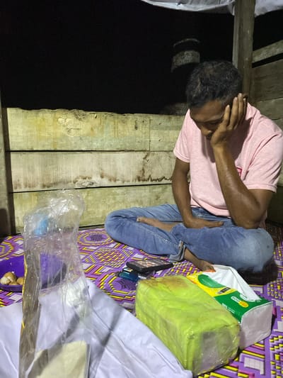 A man wearing a pink t-shirt and blue jeans sits cross-legged on a mat with a purple and yellow geometric pattern. He is leaning his head on his left hand, with his eyes downcast. In front of him, to his right, is a pack of facial tissues, partially wrapped in green plastic, next to a white tissue box. To his left, a clear plastic bag contains a few small, round, light-colored objects, possibly food. There is also a purple bowl visible. The background consists of weathered wooden planks, and the scene is lit by an artificial light source, suggesting it is nighttime. The location is described as the city of Lamtui, Indonesia.