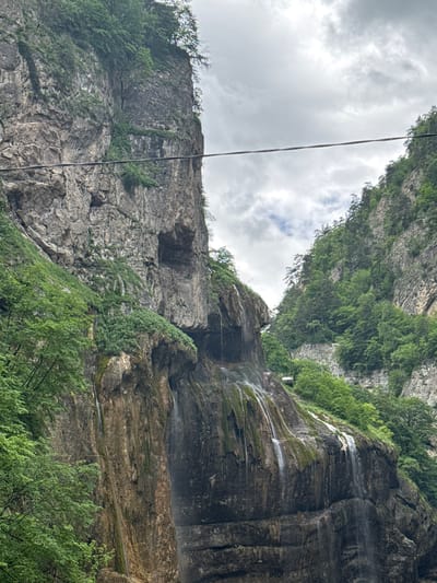 The image shows a natural landscape in Khushto-Syrt, Russia, characterized by a narrow mountain gorge with steep, rocky cliffs on either side. A waterfall cascades down the face of the left cliff, with water streams visible running over moss-covered rock. Lush green vegetation, including trees and bushes, grows densely on the slopes of the cliffs. A wire, possibly part of a power line or cable car system, stretches horizontally across the upper portion of the gorge. The sky is overcast with clouds, suggesting a diffused light. In the distance, on the right side of the gorge, a small, white structure with a red roof is visible perched on the mountainside, potentially a building or a small vehicle. The overall scene depicts a rugged and verdant natural environment within a mountainous region.