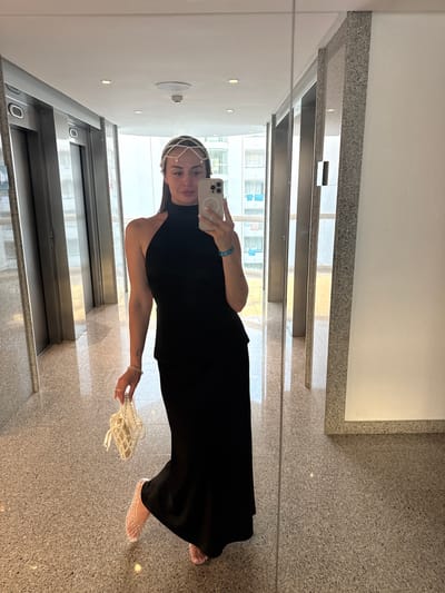 A woman stands in a hallway, reflected in a mirror. She is wearing a black, halter-neck maxi dress, a pearl headpiece, and a mesh handbag. She is holding a cell phone in her right hand, taking the photograph. Her left hand is holding the handbag, and she has a bracelet on her left wrist. She is also wearing mesh ballet flats. The hallway appears to be in a modern building, with polished granite floors and metal elevators. In the reflection, buildings with blue balconies are visible through large windows. The overall impression is one of a pre-event or post-event moment.