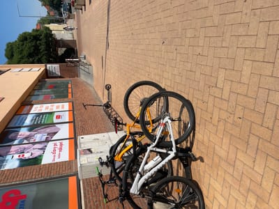 The image shows a street scene in Stupava, Slovakia, on a sunny day. The foreground is dominated by a row of parked bicycles. Two bicycles are prominently featured: one is white with the brand "ROCKRIDER" visible on its frame, and the other is orange. Several other bicycles are partially visible, some mounted on a rack. The bicycles are parked on a brick-patterned sidewalk.
Behind the bicycles, a brick building with large windows lines the street. Advertisements are displayed on the windows. One advertisement features images of an elderly couple and a smartphone, with text in Slovak that appears to promote a cooperative shopping experience. Another sign on the building reads "BIŽUTÉRIA ZLATNÍCTVO ZALOŽNÁ". Further down the street, a sign with the word "POŠTA" (Post) is visible.
In the background, trees and other buildings can be seen. A dark-colored car is parked on the street, and a person is walking away from the camera. The overall environment suggests a commercial or residential street in a European town. The perspective of the photograph is from a slightly elevated angle, looking down and along the street.