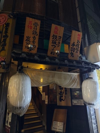 The image shows a close-up, low-angle shot of a building facade, likely a restaurant, at night. The scene is dominated by wooden signs with Japanese text, white paper lanterns covered in plastic wrap, and exposed light fixtures. The overall aesthetic suggests a traditional Japanese eatery, possibly a izakaya (Japanese pub).
The wooden signs display various Japanese characters, some in black and some in red. One sign reads "伝説お殿様の 手羽先" (Densetsu otonosama no tebasaki), which translates to "Legendary Lord's Chicken Wings." Another sign says "コリコリ 先祖鉄板つくね" (Korikori senzo teppan tsukune), potentially referring to a chewy ancestor's iron plate tsukune (meatball dish). A third sign mentions "骨付鶏もも肉の 名物 炭火烧" (Hone-tsuki dori momo niku no meibutsu sumibiyaki), indicating a signature charcoal-grilled bone-in chicken thigh.
Two large, white, cylindrical paper lanterns are prominent, both wrapped in clear plastic. One is positioned near the top right of the frame, and the other lower down and to the left. An ornate metal structure, possibly part of the building's lighting or decoration, runs vertically down the center.
A smaller white sign with Japanese text is visible near the right side, reading "私有地につき ここでの飲食、喫煙はご遠慮下さい!!! 防犯カメラ作動中" (Shiyūchi ni tsuki koko de no inshoku, kitsuen wa go-enryo kudasai!!! Bōhan kamera sadōchū), which translates to "This is private property. Please refrain from eating or smoking here!!! Security cameras are in operation." This sign provides a clear indication of rules and security measures.
The lighting is dim and atmospheric, characteristic of nighttime street scenes in Japan. The setting appears to be a narrow street or alleyway, typical of older districts. The overall mood is inviting and somewhat mysterious, hinting at a dining experience steeped in tradition.