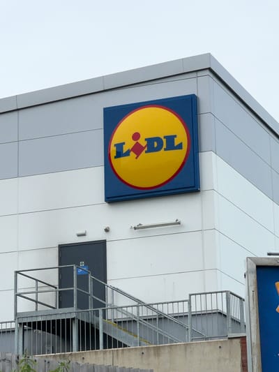 The image shows the exterior of a modern building, likely a retail store, with a LIDL logo prominently displayed. The building's facade is composed of large, light gray panels arranged diagonally, creating a contemporary architectural style. The LIDL logo itself is a circular emblem with a yellow interior and a red border, featuring the brand's name in blue letters. To the right of the logo, there's a dark gray door with a smaller blue and white sign attached.
Adjacent to the building is a metal fire escape staircase, providing access to an upper level. The staircase is constructed from galvanized steel and features a grid-like pattern for the steps and railings. To the far right of the image, part of a rougher, concrete wall is visible, suggesting a different construction material or an older section of the building. A few green leaves from a nearby plant are also visible on the right edge of the frame, indicating an outdoor setting.
The sky above is overcast and a pale gray, suggesting either early morning, late afternoon, or a cloudy day. There are no people visible in the image, and the scene appears to be static. The overall impression is that of a functional, commercial building. The presence of the LIDL logo and the context of Stoke-on-Trent, United Kingdom, strongly suggest this is a LIDL supermarket.