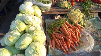 A market stall in Puloara, Indonesia, displays a bounty of fresh produce. In the foreground, a large pile of pale green cabbages is arranged on a tiered wooden structure. To the right, a vibrant mound of bright orange carrots with leafy green tops is neatly displayed. Behind the carrots, a head of cauliflower with creamy white florets rests on a weathered wooden surface, alongside more cauliflower further up and to the left. Scattered among the vegetables are red tomatoes, their skins gleaming in the sunlight, and a bundle of green beans in a plastic bag. The stall appears to be outdoors, with the wooden planks and the natural lighting suggesting a market setting.