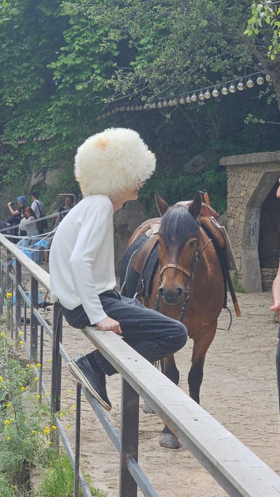 A young person wearing a fluffy white hat and a white long-sleeved shirt is perched on a wooden railing, facing away from the camera. They are wearing black jeans and dark shoes. Beside them stands a brown horse, saddled and bridled. The horse's head is turned towards the viewer, and its ears are perked. In the background, lush green trees and foliage create a natural setting. A string of lights is visible hanging from a tree branch. Other people can be seen in the distance, some taking photos. The ground appears to be dirt or sand. The overall scene suggests an outdoor recreational area, possibly a stable or park. The specific location within Красный Курган, Russia, is not determinable from the visual information.