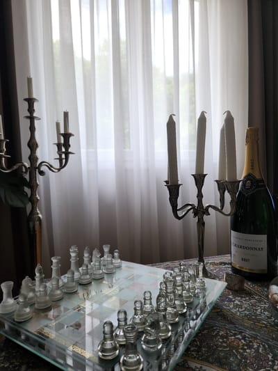 This image depicts an indoor scene located in Idar-Oberstein, Germany. In the foreground, a rectangular glass chessboard is visible, populated with two full sets of translucent glass chess pieces, one frosted and the other clear, arranged in their starting positions. The center of the chessboard features decorative text, including "Turkish Delight" and "Lokum."
To the left, a tall, ornate bronze-toned candelabra with three arms holds three unlit white taper candles. To the right, a shorter, four-armed silver-toned candelabra also holds four unlit white taper candles. Adjacent to the right candelabra stands a dark green champagne bottle, labeled "FRANÇOIS NOBLECOUR CHARDONNAY BRUT," with a gold foil cap. These items are situated on a table covered by a richly patterned fabric with intricate designs in various colors including blue, red, and gold.
The background is dominated by a large window covered with sheer, white translucent curtains, through which diffused natural light enters the room. Vague outlines of green foliage and a bright sky are discernible outside the window. No individuals are present in the image.