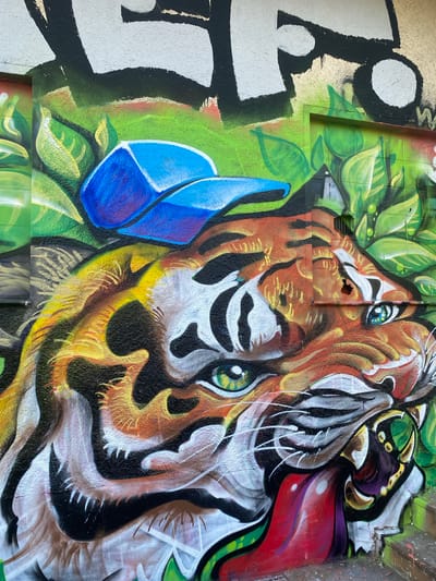 The image shows a section of graffiti art on a wall in Landau in der Pfalz, Germany. A tiger's head dominates the artwork, rendered in orange, black, white, and yellow. The tiger is surrounded by green foliage. To the left, a large black and white abstract design is painted. A blue geometric shape sits adjacent to the tiger's face. The artwork appears to be on a concrete or stone surface. A hole in the wall is visible near the tiger's head.