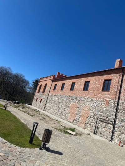 This image captures a historical building under a clear blue sky, set in what is identified as Guryevsk, Russia. The building, likely a castle or fortified structure, is constructed with a distinctive mix of materials: sections of weathered red brickwork alternate with areas of rough, irregularly shaped stone masonry. The brick sections feature multiple dark-framed rectangular windows on the upper levels, and some arched windows along with smaller rectangular openings lower down. A notable detail is the crenellated parapet visible on one part of the building, suggesting a defensive architectural style. Drainage pipes or conduits are also visible running down the facade.
In the foreground and extending to the right, the ground is a paved area comprising irregular flagstones and cobblestones, interspersed with smoother pathways and patches of bright green grass. On this paved surface, a dark-colored park bench with wooden slats, a matching cylindrical trash bin, and a modern black lamp post are visible. These objects cast long, distinct shadows, indicating a bright, sunny day, likely morning or afternoon.
Below the main building and behind the paved area, a line of deciduous trees stands without leaves, suggesting the season is late autumn, winter, or early spring. The sky above is a vast, cloudless expanse of vibrant blue. There are no people visible, and no specific activities are taking place. No discernible text is present in the image. The overall scene evokes a sense of historical presence and tranquility under clear weather conditions.