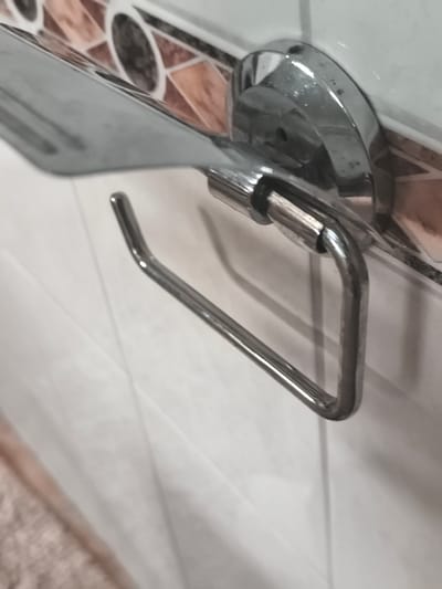 The image is a close-up, angled shot of a chrome toilet paper holder mounted on a tiled wall. The toilet paper holder has a curved arm for holding the roll and a flat shelf above it. The tiles on the wall are white and appear to be in a grid pattern. There is a decorative border of tiles with a geometric pattern above the white tiles. The lighting is bright, creating reflections on the chrome surfaces. No people are visible in the image. The scene appears to be in a bathroom.