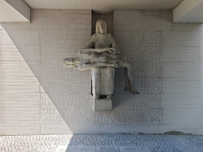 This image shows a striking memorial sculpture set against a textured concrete wall. The sculpture, carved from stone, depicts a grieving woman cradling a fallen figure in her lap, a pietà-like representation. The figures are stylized and weathered, adding to the somber mood. The setting appears to be outdoors, with a prominent shadow cast by an overhead structure, suggesting a time of day when the sun is at an angle, possibly mid-morning or late afternoon. The ground in the foreground is paved with cobblestones, indicating a public space or memorial site.
The concrete wall behind the sculpture is inscribed with numerous lines of text. To the left of the sculpture, some lines read: "MARIA KONIGINDE," "UNS DEN," "DIESES DENKMALIST," "OPFERNDES ERSTEN," "UND DEN 36,000,000," "KRIEGES 1939/1945," "DEUTSCHLAND," "FRANKREICH," "BULGARIEN," "FINNLAND," "NORWEGEN," "HOLLAND," "LUXEMBURG," "FRANKREICH," and "HERR VERGIBUNS UNSERE SCHULD." To the right of the sculpture, some visible text includes: "FRIEDENS ERFLEHE," "DEN 36,000,000 KRIEGES 1914-1918," "ZWEITEN WELT," "ENGLAND DAS," "ITALIEN," "TÜRKEI," "UNION DER KOMMUNISTISCHEN," "VEREINIGTEN VON," "AMERIKA," "JAPAN CHINA," "FRANZÖSISCHE UNION," and "SCHULD WIE AUCH." The presence of country names and references to wars, particularly "KRIEGES 1939/1945" (World War II) and "KRIEGES 1914-1918" (World War I), strongly suggests this is a memorial related to wartime sacrifices and perhaps seeking forgiveness. The text "HERR VERGIBUNS UNSERE SCHULD" translates to "Lord, forgive our guilt," reinforcing this theme. The memorial is located in Memmingen, Germany.