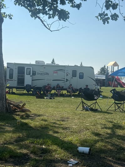 The image depicts an outdoor scene at a campsite in Lyalta, Alberta, Canada. A large white recreational vehicle (RV), labeled "SPORTSMEN," is parked in the center, with its door open. A "FOR SALE" sign is visible on the side of the RV. Several people are seated in lawn chairs around the RV. To the left of the RV, a pile of firewood is stacked near a tree. In the foreground, the grass is green and somewhat dry in patches. Several empty lawn chairs are scattered around. A blue canopy is visible on the right side of the frame, with a red slide and playground structure in the background. A single white can, identified as "Coors Light," sits on the grass to the left. Further to the right, a white tumbler and some crumpled foil are on the ground. The sky is clear and blue, with branches of a tree hanging into the frame from the top. The overall impression is of a casual, relaxed outdoor gathering at a campsite.
