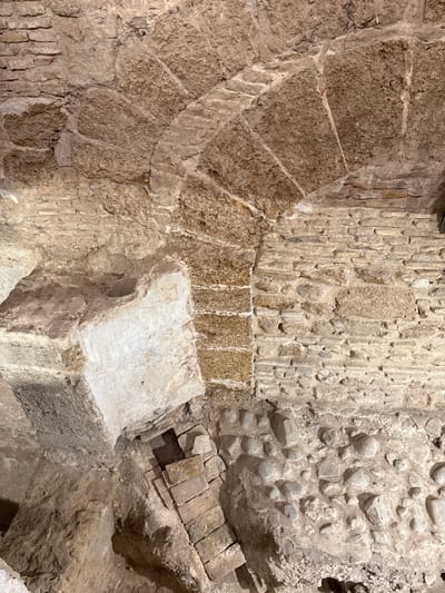 The image displays an excavated archaeological interior, specifically at the Roman Theater of Cádiz, Spain. A large, semicircular arch constructed from textured, light-brown, rough-hewn stone voussoirs dominates the upper portion. Adjacent wall sections are built with smaller, irregular stones and horizontal courses of brick, indicating diverse construction. Below, a prominent, lighter-colored block-like structure projects from the left. The excavated ground reveals a descending series of rectangular stone blocks, possibly steps, on the left, and compacted earth interspersed with numerous rounded river stones or cobbles on the right. The scene is illuminated by artificial light, emphasizing the varied textures and historical layers of masonry. No individuals or active elements are present.