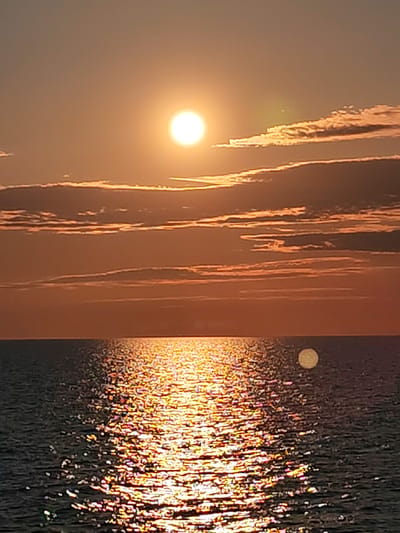 A vertical, eye-level, medium shot captures a sunset over the sea. The sun, a bright orb, is positioned in the upper left quadrant of the image, casting a warm orange glow across the scene. Long, horizontal clouds, tinged with pink and orange, stretch across the sky. The sea below is a deep blue, with a shimmering reflection of the sun creating a dazzling path of light across its surface. In the distance, a faint, circular object, possibly the moon, is visible in the sky to the right of the sun. The scene is serene and tranquil, evoking a sense of peace and beauty. The image was taken in Kakkarovo, Russia.