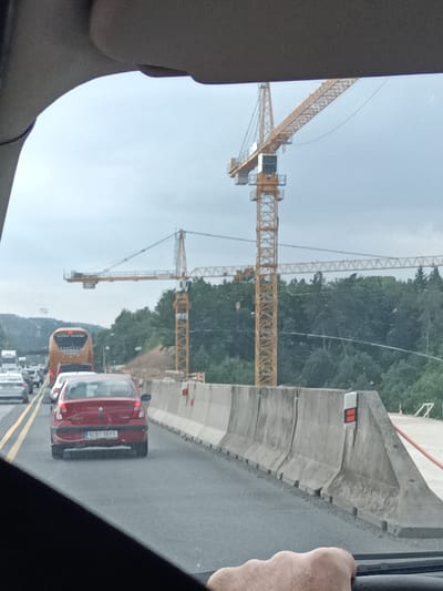 The image shows a view from inside a vehicle looking forward at a highway construction site in Hrusice, Czechia. Two large yellow tower cranes are prominent in the scene, with one in the foreground and another further back. The cranes are positioned above a section of the highway that is undergoing construction, indicated by concrete barriers lining the road and piles of earth.
The highway itself appears to be congested, with several cars and a bus visible in traffic. The red car in the foreground has the license plate "5C8 3911". The sky is overcast, suggesting a cloudy or possibly rainy day. The surrounding area is characterized by dense green trees and a distant hilly landscape. The overall atmosphere appears to be one of a typical day on a busy road with ongoing infrastructure work.