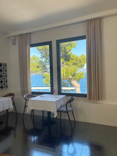 This image depicts a restaurant interior with a view of the Adriatic Sea in Korčula, Croatia. Two tables are set with white tablecloths and chairs. The table in the foreground has two chairs, one on each side, and is adorned with a small vase containing a pink flower and two glasses. The second table, partially visible on the left, is also covered with a white tablecloth. Two large windows dominate the background, offering a scenic vista of the sea, a large pine tree with a curved trunk, and a clear blue sky. The floor is tiled with large, dark, reflective squares. Beige curtains hang on either side of the windows. A decorative tile panel with a white floral pattern on a dark background is mounted on the left wall. A white speaker is visible on the upper left wall, and a circular smoke detector is mounted on the ceiling. The overall atmosphere suggests a serene dining setting with a focus on the natural beauty of the coastal landscape.