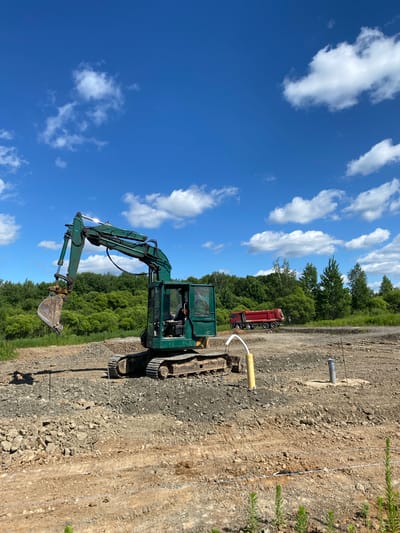 An outdoor, eye-level, medium shot captures a construction site under a bright, partly cloudy blue sky. In the foreground, a green excavator sits on a mound of dirt and gravel, its boom extended and bucket lowered. To the right of the excavator, a yellow pipe with a white tube curving out of it stands vertically, and further to the right, a gray pipe emerges from a concrete base. In the middle ground, a red dump truck is parked on a dirt road. The background consists of a dense line of green trees. The ground is a mixture of loose dirt, gravel, and some larger rocks. There are no visible people.