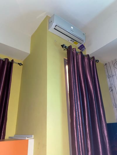 The image depicts an indoor room located in the city of Entalsewu, Indonesia, viewed from a low angle looking upwards. The walls are painted a pale yellow-green, meeting a white ceiling. A white Sharp split-unit air conditioner is mounted high on the wall near the ceiling, displaying "26" on its digital screen and bearing an SNI (Indonesian National Standard) label. Directly below the air conditioner, a small, crumpled piece of purple material, possibly packaging, is affixed to the wall, partially obscured by a curtain rod.
Two dark purple, satiny eyelet curtains are visible, hanging from silver-colored rods with decorative black and white finials. The curtains are drawn, covering what appears to be a window or windows, with a sliver of light visible from behind the right curtain. In the bottom left corner, a portion of orange furniture, possibly a desk or cabinet, is visible, topped with white objects. The far right section of the wall features a white surface with subtle grey patterning, resembling wallpaper. Minor scuff marks and discoloration are present on the yellow-green walls. No individuals are present in the scene.