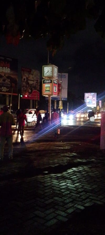 The image displays an urban street scene at night in Tudor ward, Kenya. The foreground reveals a wet, herringbone-patterned brick pavement, reflecting ambient light. Centrally located in the mid-ground is a street clock tower featuring a square clock face, indicating approximately 10:10. Below the clock, illuminated advertising panels for the "MIKA" brand are visible. To the left, a large billboard partially displays "Wild Sale" and "50%," positioned near a red traffic light. The road in the mid-ground is visibly wet, reflecting bright headlights from multiple vehicles, which cause significant lens flare across the frame.
Several individuals are present: a person in light-colored pants and a darker upper garment is in the left foreground, partially obscured. Closer to the roadway, multiple figures are discernable, including one wearing a red shirt, another on a bicycle, and additional indistinct pedestrians. A white car is observed moving rightward on the road. Further billboards with illuminated advertisements are visible in the background against the dark night sky. Dark foliage is present at the top of the frame.