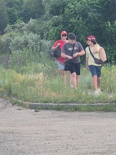A medium shot captures three individuals walking on a dirt path alongside a grassy embankment. The person on the left, wearing a red t-shirt and a red cap, carries a black bag. In the center, a person in a gray t-shirt with the letters "M/D" printed on it, and black shorts, walks with a black strap across their body. To the right, a person in a yellow t-shirt and blue shorts, also wearing a red cap, carries a tan bag slung over their shoulder. The background is filled with lush green trees and vegetation. The scene appears to be outdoors, possibly in a park or natural area. The image does not provide specific details about the city of Pidhaitsi, Ukraine.