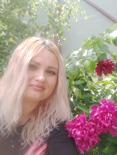 A woman with blonde hair is taking a selfie in a garden. She is wearing a black shirt. In the background, there are pink peonies and green leaves. The location is in the city of Изыхские Копи, Russia.