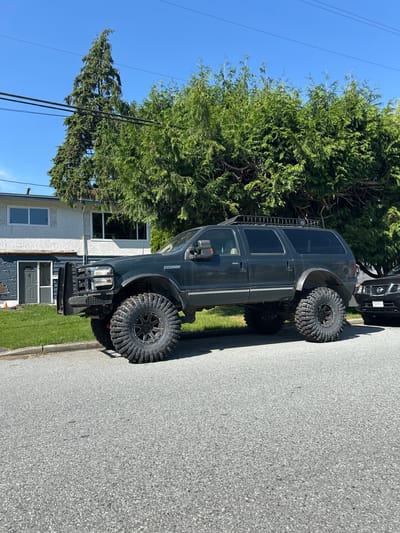 A dark-colored Ford Excursion SUV is parked on the right side of a residential street in Port Coquitlam, Canada. The SUV has oversized tires, a roof rack with a ladder, and a brush guard on the front. A portion of a black Nissan vehicle is visible parked behind the SUV. To the left of the SUV is a two-story house with white siding and blue trim. A large green tree is behind the house, obscuring the top portion. Utility poles and lines are visible in the blue sky. The road is paved and slightly wet or reflecting light. A small patch of green grass is visible between the road and the house. The time of day appears to be midday under clear conditions, indicated by the shadows and the bright blue sky. The scene is set in a residential neighborhood.