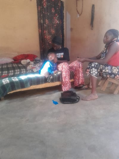 Inside a simple room, three people are visible. A woman in a coral top and floral skirt sits on a stool near the wall. On a bed or padded bench, a child lies horizontally. A person in a black t-shirt with white and red lettering stands over the child. A black bag is on the floor next to the bench. The room has a rustic feel with plain walls and a floral fabric hanging on one side.