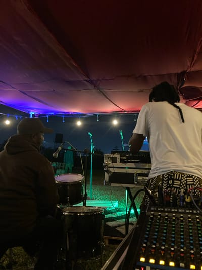 An outdoor music performance setup is depicted at night in Murera ward, Kenya. A dark fabric canopy overhead is illuminated by string lights and colored stage lighting, exhibiting blue on the left and red on the right. On the left, a person wearing a brown hoodie and a baseball cap is seated, facing right, positioned in front of at least two drum kit components. On the right, a second individual with dreadlocks, wearing a white t-shirt and patterned shorts, stands, actively operating rack-mounted audio equipment on a stand. In the immediate foreground, a multi-channel audio mixing console with illuminated controls is visible. The ground is a grassy surface, partially illuminated by green stage lighting. Several microphones on stands are positioned center-background. Behind the drum setup, to the left of the microphones, several chairs and a faintly illuminated, possibly mannequin-like, figure are present. The background shows a dark night sky above the horizon.