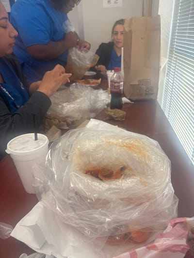 The image captures an indoor scene in Grand Prairie, Texas. A group of people are gathered around a table. One person wearing a blue shirt can be seen on the left side, holding a bag of food, another person wearing a black shirt with blue sleeves is seen further down. A woman with dark hair is sitting to the upper right of this person wearing a blue shirt, she is looking at the camera. 

On the table are numerous clear plastic bags filled with food, likely seafood. There is a large white paper cup with a straw, a brown paper bag, and a bottle of Coca-Cola. The environment appears to be a casual dining or break room setting, with the blinds in the background indicating the presence of a window.