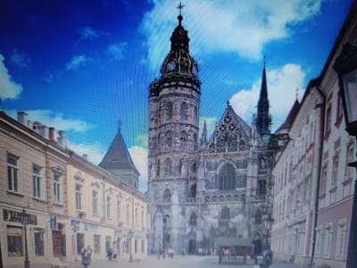 The image captures a street view in the city of Košice, Slovakia. In the center, St. Elizabeth's Cathedral, a prominent Gothic church, dominates the scene with its intricate architecture and tall spire topped with a cross. To the left of the cathedral, a series of light-colored buildings with traditional architectural features line the street, featuring multiple windows and balconies. A street lamp with a sign that reads "PAR" is visible in front of one of these buildings. On the right side of the image, another building with a pink facade and white trim stands. The street is paved, and in the foreground, a few people are present, some walking and others appearing to be interacting. The sky is a vibrant blue with white clouds, suggesting fair weather. The overall scene depicts a historic urban setting with significant architectural landmarks.