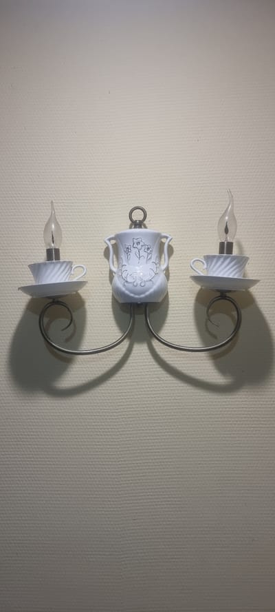 This media file shows a vertically oriented wall sconce mounted on a textured, light-colored wall. The sconce is designed to resemble three candle holders made from white porcelain teacups. Each teacup is oriented upside down, with its opening facing downwards.
The top and bottom elements of the sconce feature a clear, flame-shaped glass bulb with visible filaments, simulating a candle. These bulbs are held within the teacup, with the saucer part of the teacup acting as a shade. The middle element is a teacup with a decorative floral pattern in a silver or gray color. This teacup also has two small handles.
The teacups are attached to a curved, dark metal frame that supports them in a vertical arrangement. The frame has a decorative spiral element. The entire sconce casts a shadow on the wall to the right.
There are no people visible in the media file. The scene appears to be indoors, with artificial lighting contributing to the overall illumination and the shadow casting. The wall's texture suggests it might be wallpapered. No text is visible. The overall impression is of a decorative lighting fixture with a vintage or whimsical aesthetic. The location context provided is Chernomorskiy, Russia, which does not offer specific visual cues within the image itself.