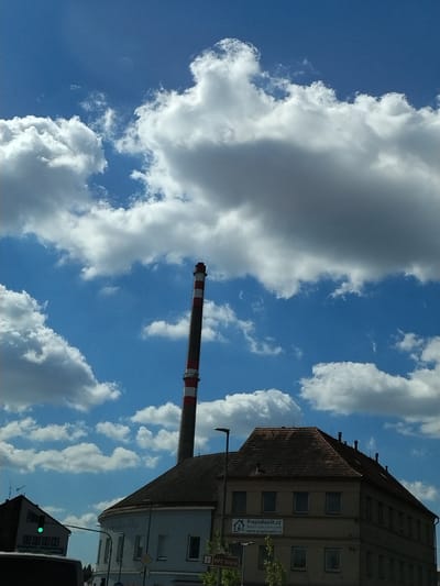 The image shows a street scene in Strakonice, Czechia. A tall, red and white striped smokestack dominates the center-left of the frame, rising into a bright blue sky filled with fluffy white clouds. In the foreground, a light-colored, multi-story building with a dark, tiled roof is visible on the right side of the image. The building has numerous windows and appears to be an office or commercial property. A sign on the building reads "PrepisRealit.cz" with a website address below. To the left of this building, a darker, angular building with a green traffic light is partially visible. Trees and streetlights are also present. The overall impression is a sunny day in an industrial or commercial area.