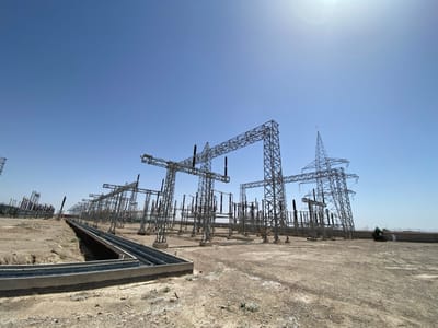 The image shows a large electrical substation, possibly a power plant, in Pir Dust, Afghanistan, a city located at coordinates 31.633, 65.854. The substation is composed of numerous steel lattice towers, connected by high-voltage cables. The towers are tall and slender, with multiple crossbars and insulators, and some of the towers are connected to a ground-level concrete structure. This structure appears to be a transformer bank. The image is taken from the ground level, facing the substation. In the distance, there is a large open area with a low dirt embankment. A small, unidentified vehicle and a single person are visible in the distance, moving towards the right side of the image. The sky is clear and blue, with the sun shining brightly. The ground is covered with dirt and rocks. The image is likely captured during the day, as indicated by the bright sunlight and the shadows cast by the towers.