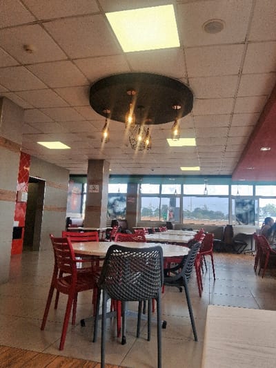 This image depicts the interior of a dining establishment, possibly a restaurant or food court, located in Kahawa Sukari ward, Kenya. The scene is largely unoccupied, viewed from a slightly elevated position looking towards large windows at the rear.
The ceiling features a grid of light-colored acoustic tiles, with several rectangular, illuminated LED panel lights. A prominent circular black decorative fixture hangs centrally, adorned with multiple illuminated Edison-style bulbs encased in dark metallic cages. A white circular speaker and a smoke detector are also visible on the ceiling.
The flooring consists of light grey tiles, with a section of wood-patterned tiles in the lower left foreground.
Multiple empty circular tables with light-colored (likely white or light wood) tabletops and metal bases are arranged throughout the space. A majority of the chairs are red plastic or painted metal, featuring slatted backs and seats. In the immediate foreground, a dark grey chair with a mesh-patterned back and seat is positioned at a table.
To the left, a structural pillar exhibits a red and white geometric tile pattern. Grey structural pillars are present further back, one displaying a "No Smoking" symbol. The rear wall is dominated by large windows, providing a view of an outdoor area with distant trees and what appears to be a road or open space.
On the far right, a single individual is partially visible from the back, seated at a table. Their head and upper torso are discernible. The establishment's overall appearance suggests a modern, casual dining environment.