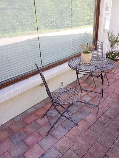 An outdoor scene in Brannenburg, Germany, features a wrought-iron patio set with a round table and two chairs placed on a brick patio. A small potted plant sits on the table. Behind the patio set, a large window is covered with white pleated blinds. A small tree with greenery and a potted plant with pink flowers are visible to the right of the patio set. A white mailbox is mounted on the wall to the right of the window. The overall environment suggests a residential or commercial outdoor space.