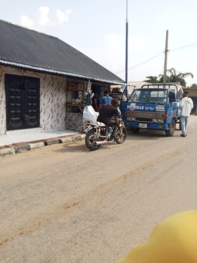 The scene is a street in Sapele, Nigeria, at coordinates 5.871, 5.691. A blue truck is parked on the right side of the street. There is a man in a white shirt standing next to the truck. A man on a motorcycle is driving toward the left. There is a small building with a black door on the left side of the street. The building has a corrugated metal roof. There are other people in the scene, but they are not visible. The street is paved with asphalt. The sky is clear and blue, with a few white clouds. There are trees and power lines in the background. The truck has writing on the side that says "DIAMONDMAN Table Mate" and "TOYOTA". The motorcycle is black and has a silver exhaust pipe. The building has a white tiled facade and is adorned with decorative tiles in a pattern of white and brown. There are some items in the window of the store. There is a gray concrete curb in front of the building. There is a man in a gray shirt on the motorcycle, and he is wearing a helmet. The truck has a black bumper and black side steps. The ground is dry and dusty. The scene is typical of a rural town in Nigeria.