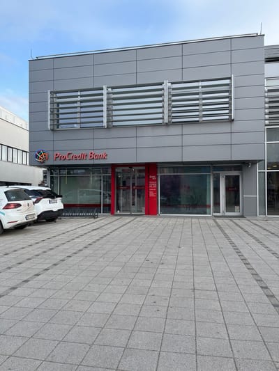 The image depicts the exterior of a modern commercial building located at Obrenovićeva Street 10 in Niš, Serbia, identified as a ProCredit Bank branch. The building features a grey panelized facade. The ground floor houses the bank, distinguished by "ProCredit Bank" in red lettering and a multicolored logo above the entrance. The entrance consists of red-framed glass automatic doors. To the right of the entrance, a red panel displays "Zona 24/7" and "OTVORENO NON-STOP" in white Serbian Cyrillic text, indicating a 24/7 access zone. Large display windows are present on the ground floor. The upper story features multiple rectangular windows fitted with horizontal silver louvers.
In the foreground, a wide area of grey rectangular paving stones extends from the building, incorporating darker grey linear patterns. Two vehicles are parked on the paved area to the left: a white Volkswagen Golf-type hatchback with partially visible Serbian license plate "NI 002-VV" and white lettering on its side, and a white SUV partially visible behind it, showing a "BG" license plate prefix. A silver metal bicycle rack is positioned on the pavement in front of the bank entrance. A small security camera is mounted on the building's facade above the right display window. The sky is partially cloudy with a light blue tone. No individuals are present in the scene.