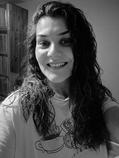 A medium shot, black and white, selfie shows a young woman with long, wet, wavy dark hair. She is smiling broadly, showing her teeth, and looking directly at the camera. Her hair is parted on the side and falls down around her face and shoulders. She is wearing a t-shirt with a graphic of a steaming coffee cup on it. In the background, to the left, is a wooden cabinet or wardrobe. The lighting is bright, creating highlights on her skin and hair. The setting appears to be indoors. There is no discernible location in Iskrets, Bulgaria.