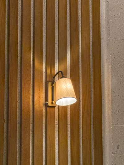 A wall sconce illuminates a wall with alternating horizontal bands of brown wood and light gray stone. The sconce has a conical shade of light beige material with a black gooseneck arm and a rectangular bronze base. The wood bands are evenly spaced and display a subtle grain. The stone bands appear rough and textured. The lighting from the sconce casts a warm glow. The location is not evident from the image.
