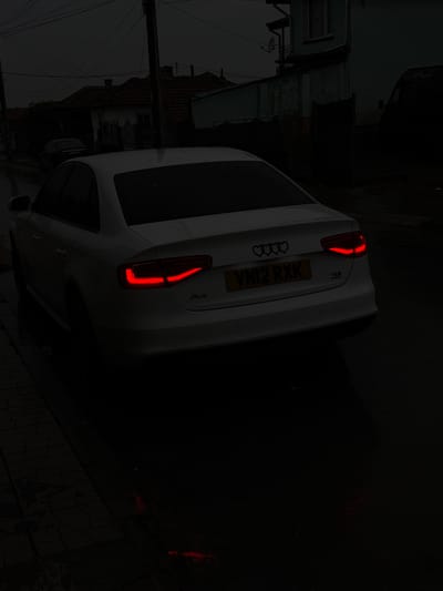 The image shows the rear of a white Audi A4 parked on a street in Dolni Tsibar, Bulgaria. The vehicle's taillights are illuminated. The registration plate is visible and reads VN12 RXK. The surrounding environment appears dark and wet. Buildings are visible in the background, but details are obscured by the darkness. A utility pole is present on the left side of the image. The focus is primarily on the car and its features.