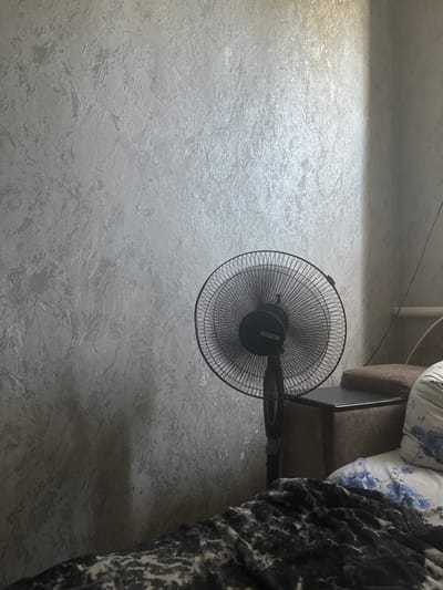 An indoor scene depicts a black oscillating fan positioned on a dark side table next to a brown upholstered chair. The wall behind the fan and chair has a textured, light grey wallpaper. In the foreground, a dark blanket with a grey and white patterned design lies on a surface, possibly a bed. A pillow with a blue floral pattern is visible on the chair. The lighting suggests artificial illumination from the left, casting a shadow of the fan onto the wall. The location is a room in Girey, Russia.