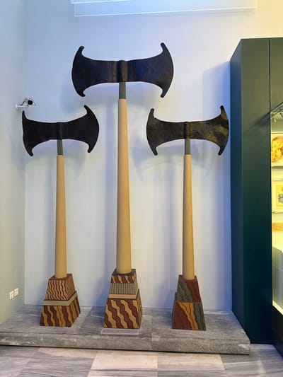 In Heraklion, Greece, within a museum setting, three imposing double-bladed axes are displayed. The axes, each with a dark, textured blade, are mounted on light-colored, cylindrical shafts. These shafts rise from decorative, tiered bases that feature geometric patterns in brown, red, and yellow. The axes are arranged from left to right in decreasing height, with the tallest in the center. They stand on a grey, stone platform that extends across the foreground. The background is a plain, light blue wall, with a security camera visible on the left. To the right, a dark green display cabinet with glass fronts offers a glimpse of artifacts within, including what appears to be pottery or stone objects. The floor is tiled with a marble-like pattern.
