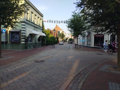 The image depicts a street scene in Cuxhaven, Germany, specifically on the Lohse-Weg, near the intersection with Kaemmererplatz. A light-colored sedan is parked in the middle of the street. To the left, a light green building features a sign for "Schleyer Versicherungen" and a blue and white traffic sign indicating a pedestrian and bicycle path. White awnings extend from the building's storefront. Further down the street, a white building on the right houses a "Fahrradgeschäft" (bicycle shop). Two women are riding bicycles towards the camera, one in a blue and white striped shirt and the other in a pink checkered jacket. Banners displaying latitude and longitude coordinates, "X 53° 5' 2' 12. 6'' N", are strung across the street overhead. The street is paved with cobblestones and brickwork, and trees line the sidewalks, casting shadows from the sunlight. The sky is clear and blue.