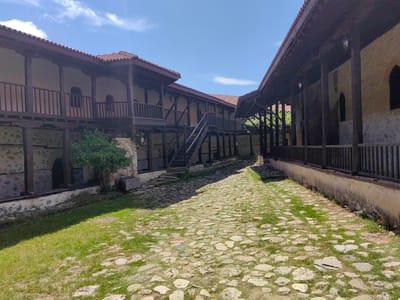 This image captures a sunlit courtyard within a historic architectural complex, identified as being in Rozhen, Bulgaria. The scene is dominated by two-story buildings featuring traditional wooden architecture. On both the left and right, overhanging upper floors with wooden balustrades and supporting posts create covered walkways or balconies, topped with distinctive red-tiled roofs. The lower sections of the buildings are constructed from stone and plaster, with some exhibiting arched windows or doorways.
A winding cobblestone path, interspersed with patches of lush green grass, stretches from the foreground towards the background, creating a sense of depth. Sunlight casts shadows from the structures and trees, highlighting the texture of the stones and grass. The sky above is a clear, vibrant blue with a few scattered white clouds, indicating a bright and sunny day.
No people are visible, and no specific activity is taking place, contributing to a tranquil and serene atmosphere. Small plants and grass grow between the cobblestones and along the base of the walls. On the right-hand structure, several wall-mounted lanterns can be seen. The architectural style, with its heavy timber frames and stone foundations, suggests a traditional monastery or an old fortified complex. No discernible text is visible within the image.