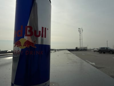 The image depicts a Red Bull energy drink can positioned in the foreground, specifically on a light grey, textured surface, likely a concrete parapet or ledge. The can is upright, prominently displaying its red, silver, and blue branding, including the "Red Bull" text and the two bull logo with "ENERGY DRINK" beneath.
In the mid-ground and background, a paved area, consistent with a rooftop parking lot, is visible. White directional arrows are painted on the asphalt surface. A dark-colored SUV is present on the right side of the paved area, with another lighter vehicle partially visible behind it. A tall, dark, lattice-style communication or cellular tower stands against the horizon. Other indistinct structures are visible in the distance. The sky is overcast and uniformly bright, suggesting a cloudy daylight condition. The scene is located in Naaldwijk, Netherlands, consistent with a commercial or industrial zone within the city. No individuals are present, and no active interactions or specific actions are occurring.